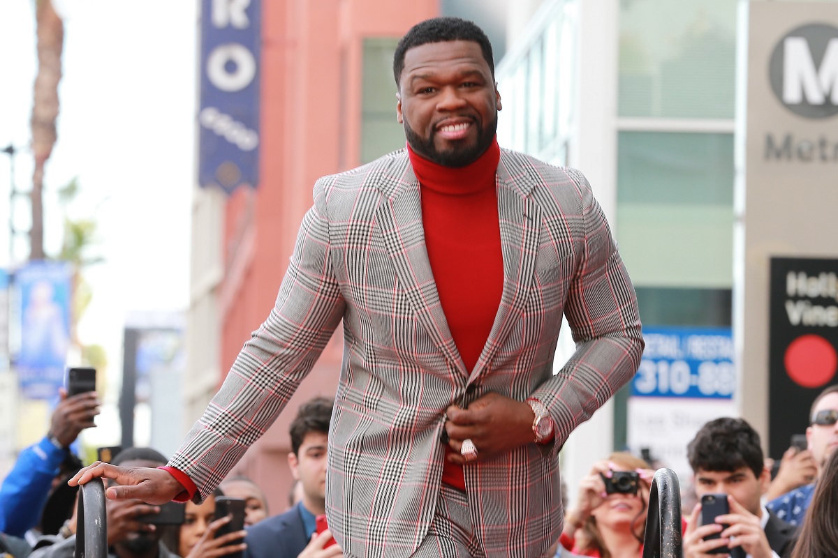 50 Cent sourit en portant un col roulé rouge et un blazer gris.