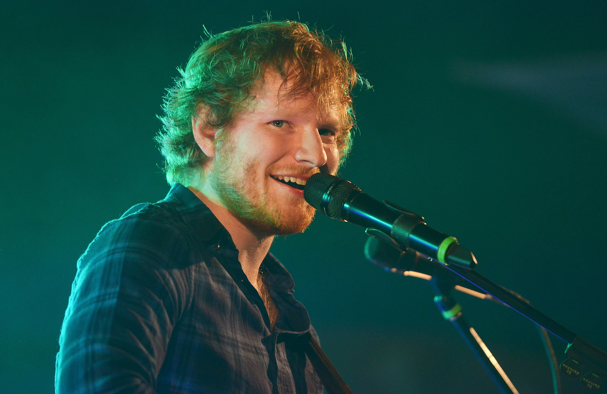 Ed Sheeran sourit en chantant dans un micro sur scène.