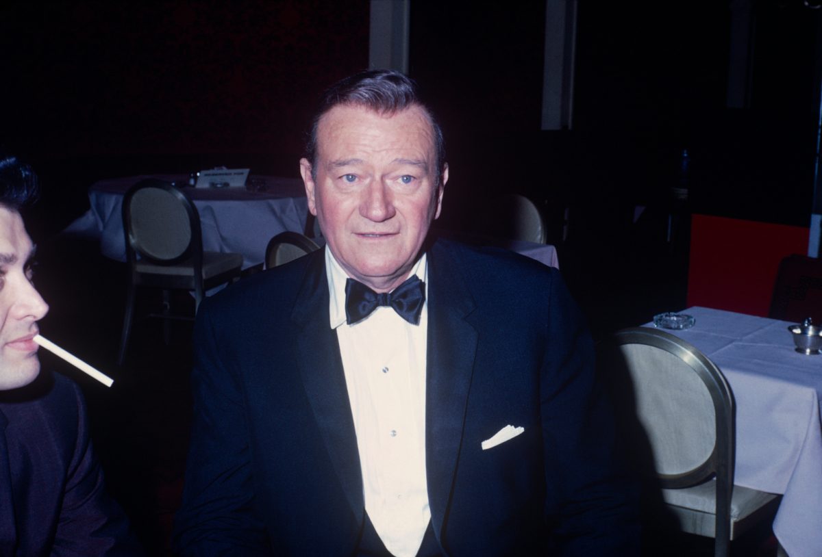 John Wayne, qui avait des opinions négatives sur la célébration des droits des homosexuels, vêtu d'un smoking noir et blanc avec des tables recouvertes de nappes blanches en arrière-plan