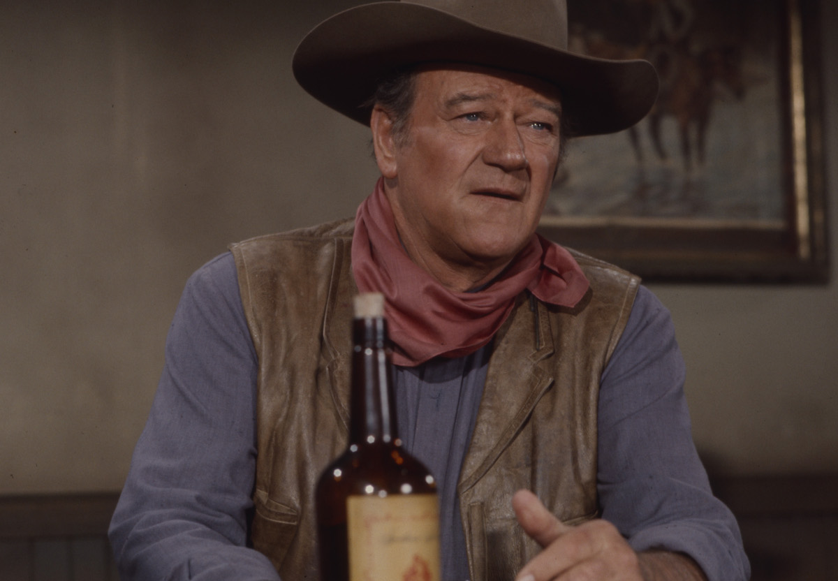 John Wayne est assis dans un saloon western avec une bouteille de whisky