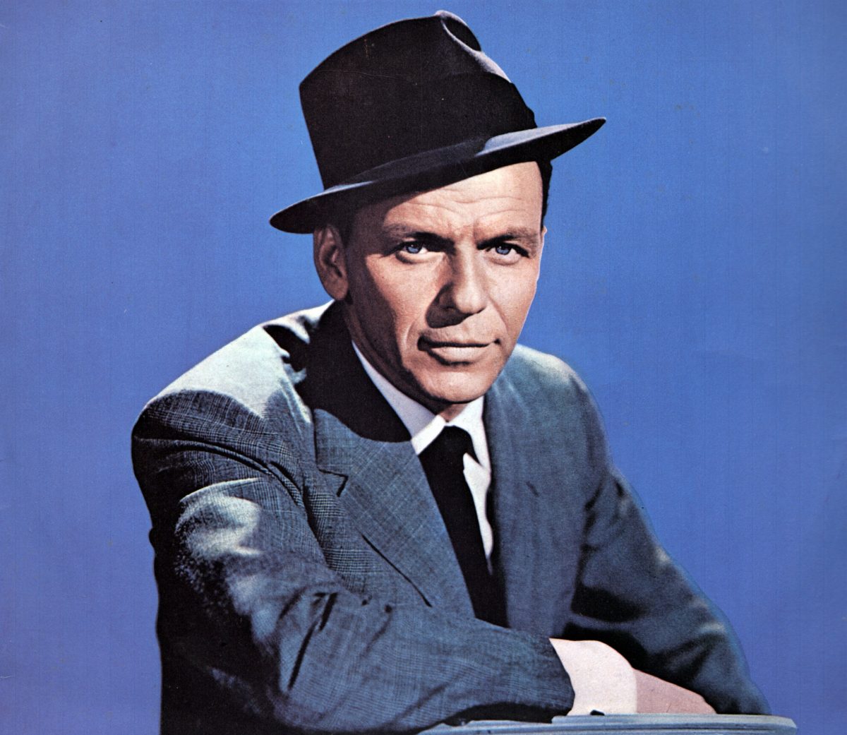 Frank Sinatra porte un chapeau et un costume et est assis devant un fond bleu.