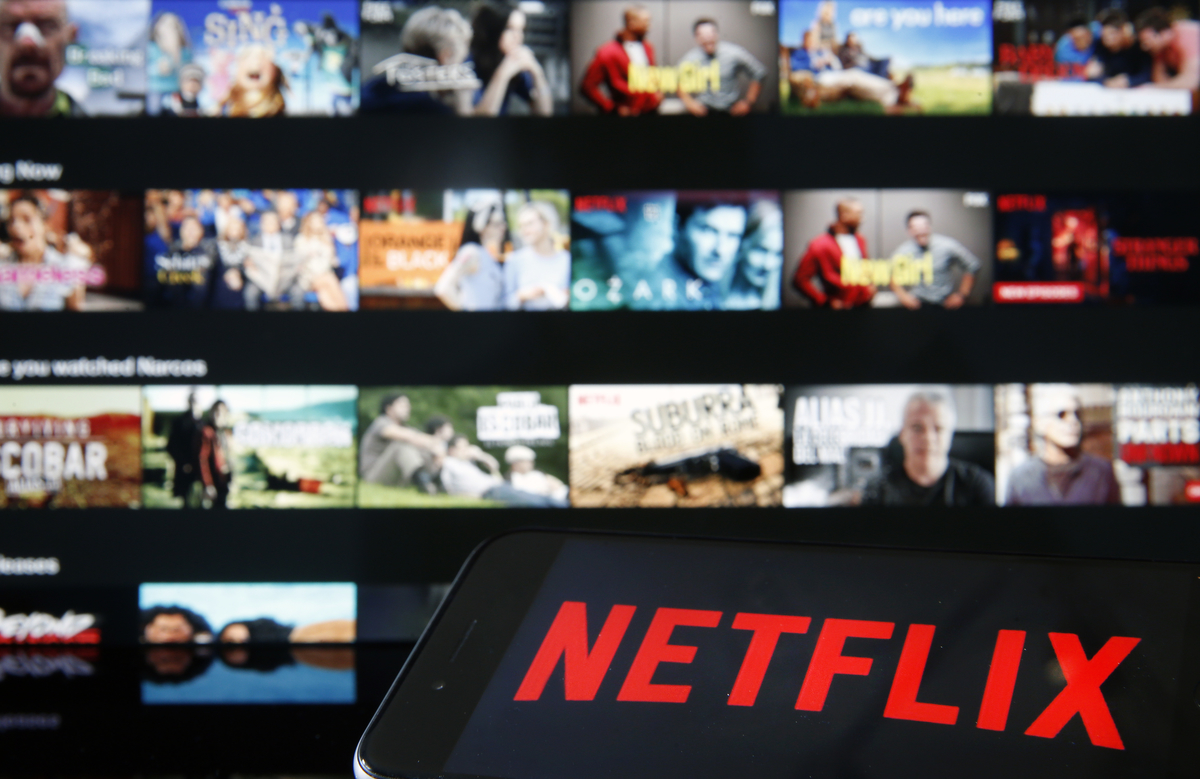 Logo Netflix sur un ipad devant un écran de télévision rempli de listes de films de Netflix.