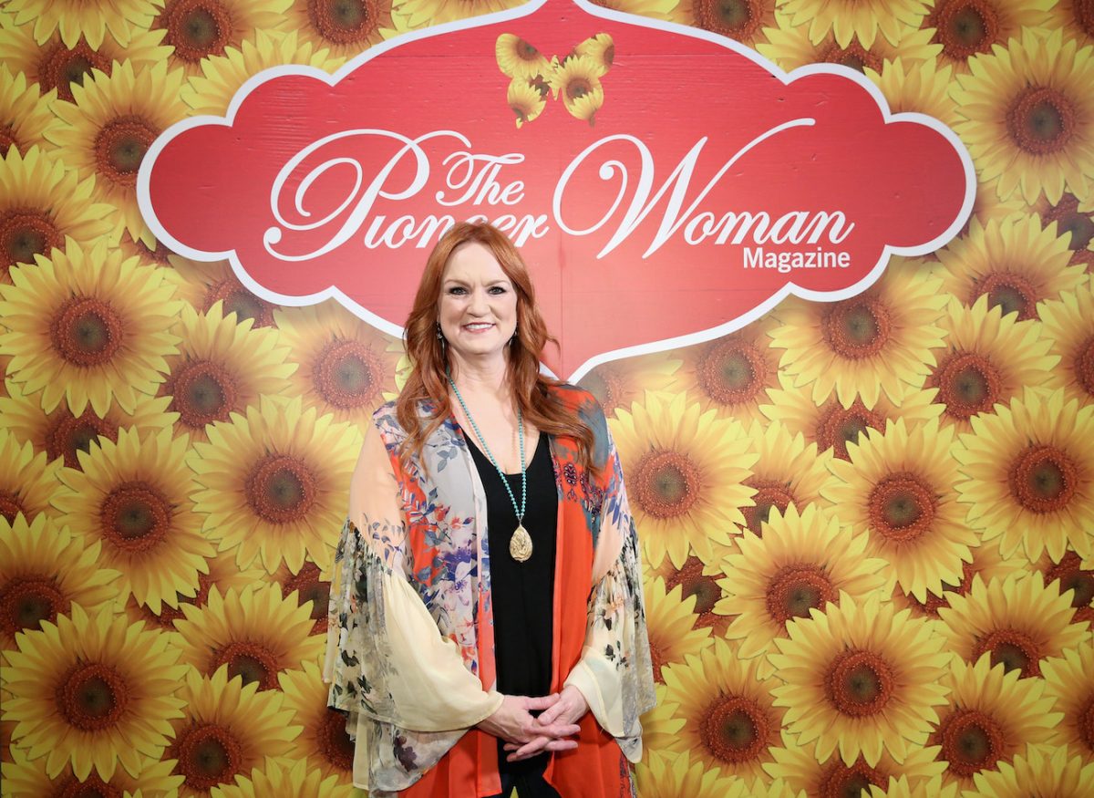 Ree Drummond sourit et porte un haut brillant fluide tout en se tenant devant un mur de tournesol jaune