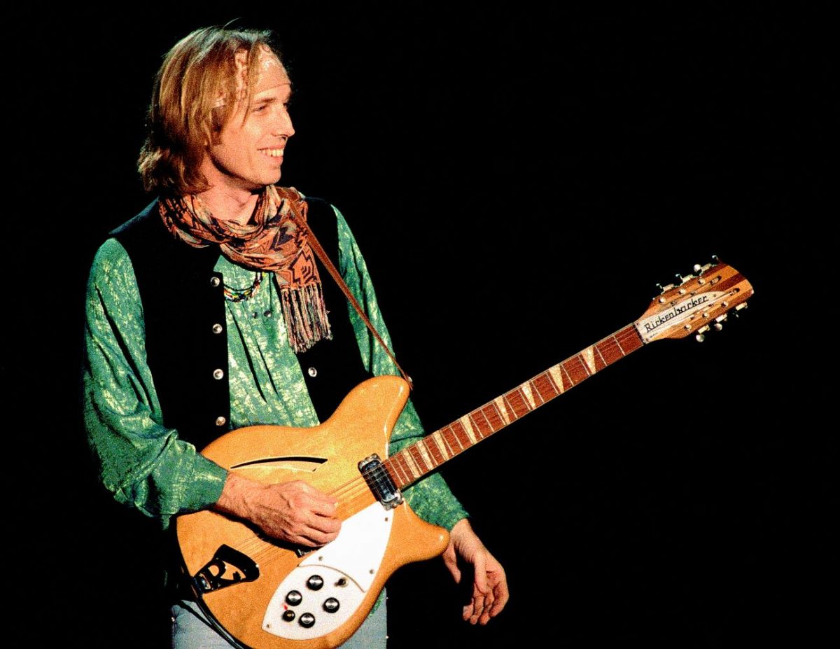 Tom Petty porte une chemise et une écharpe vertes et tient une guitare.  La biographie de Tom Petty détaille son ascension vers la gloire.