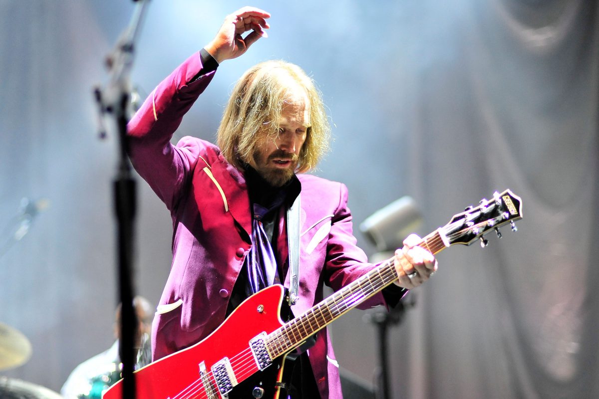 Tom Petty porte une veste violette et tient sa main en l'air tout en jouant de la guitare.