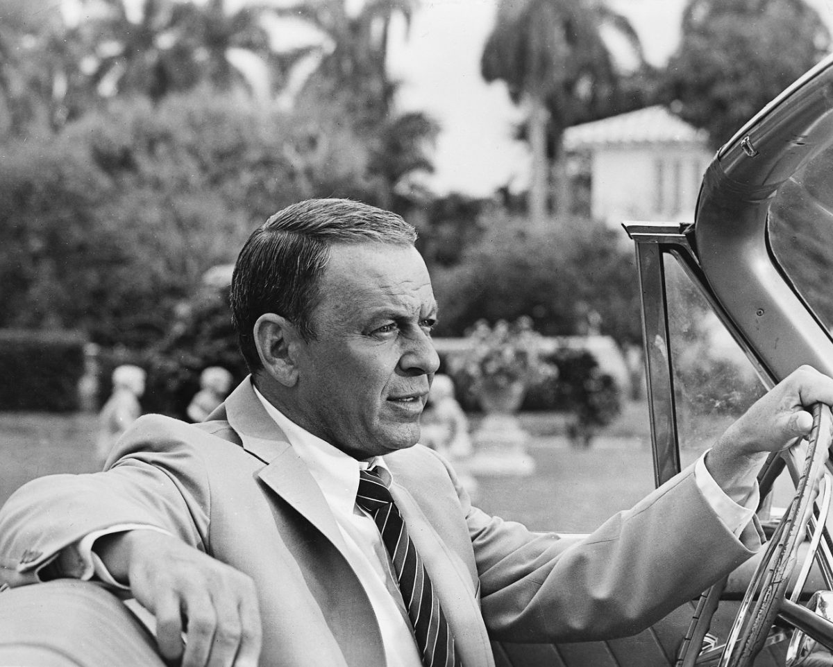 Le chanteur et acteur Frank Sinatra porte un costume et est assis au volant d'un cabriolet.