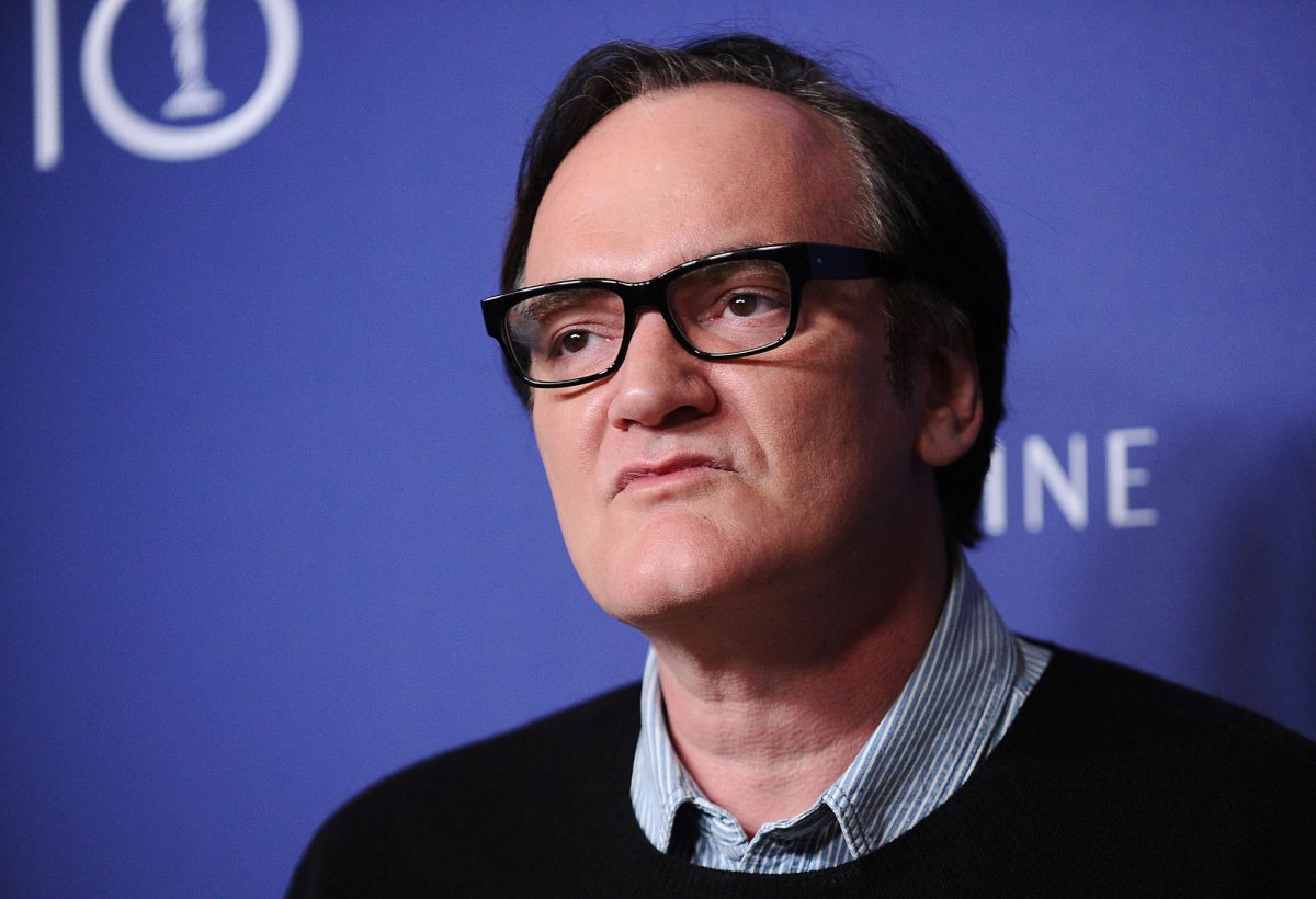 Quentin Tarantino, qui collabore avec des acteurs récurrents, portant des lunettes et une chemise à col devant un step and repeat bleu