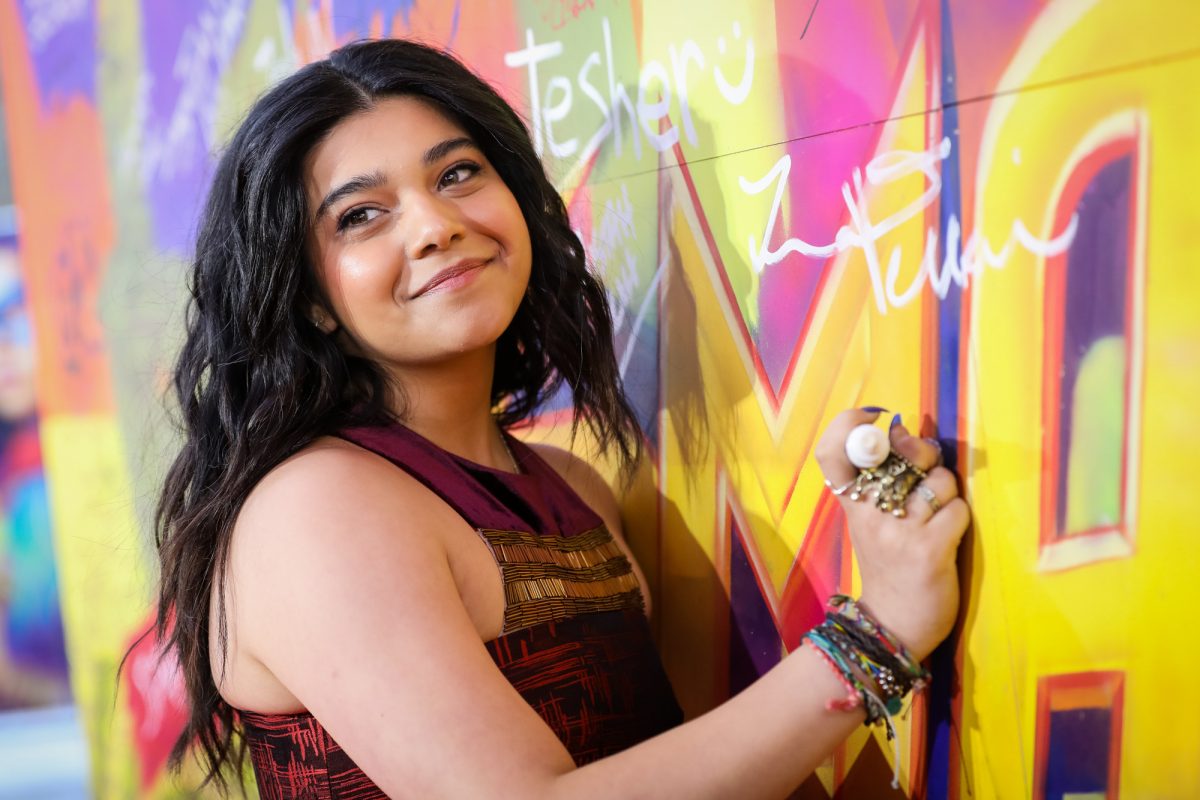 'Mme. La star de Marvel, Iman Vellani, lors de la projection de l'émission Disney+. Elle porte une robe rouge et noire avec de l'or sur le devant et elle est appuyée contre un mur aux couleurs vives.