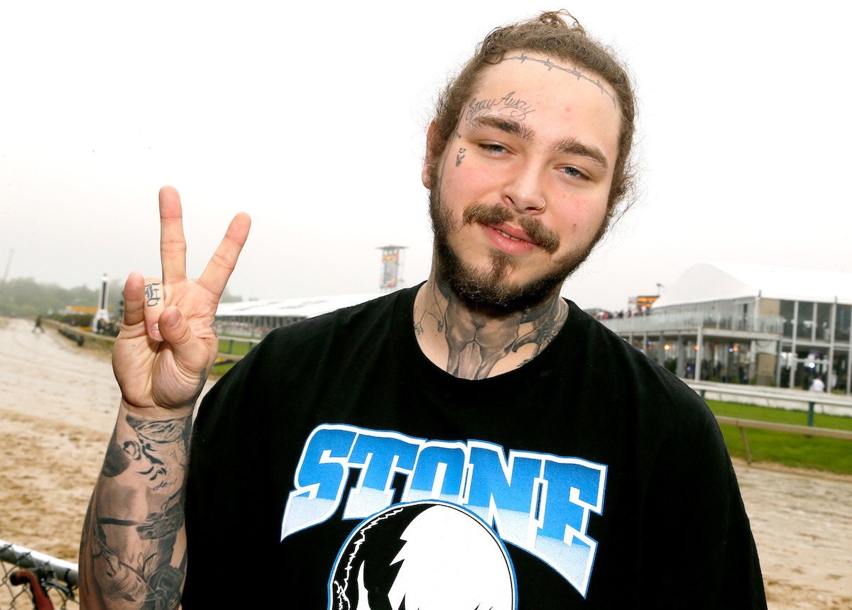 Post Malone souriant et tenant un signe de paix avec ses doigts tout en se tenant à l'extérieur. Post Malone et sa fiancée font l'objet de beaucoup d'attention depuis qu'il a annoncé la naissance de sa petite fille en 2022.