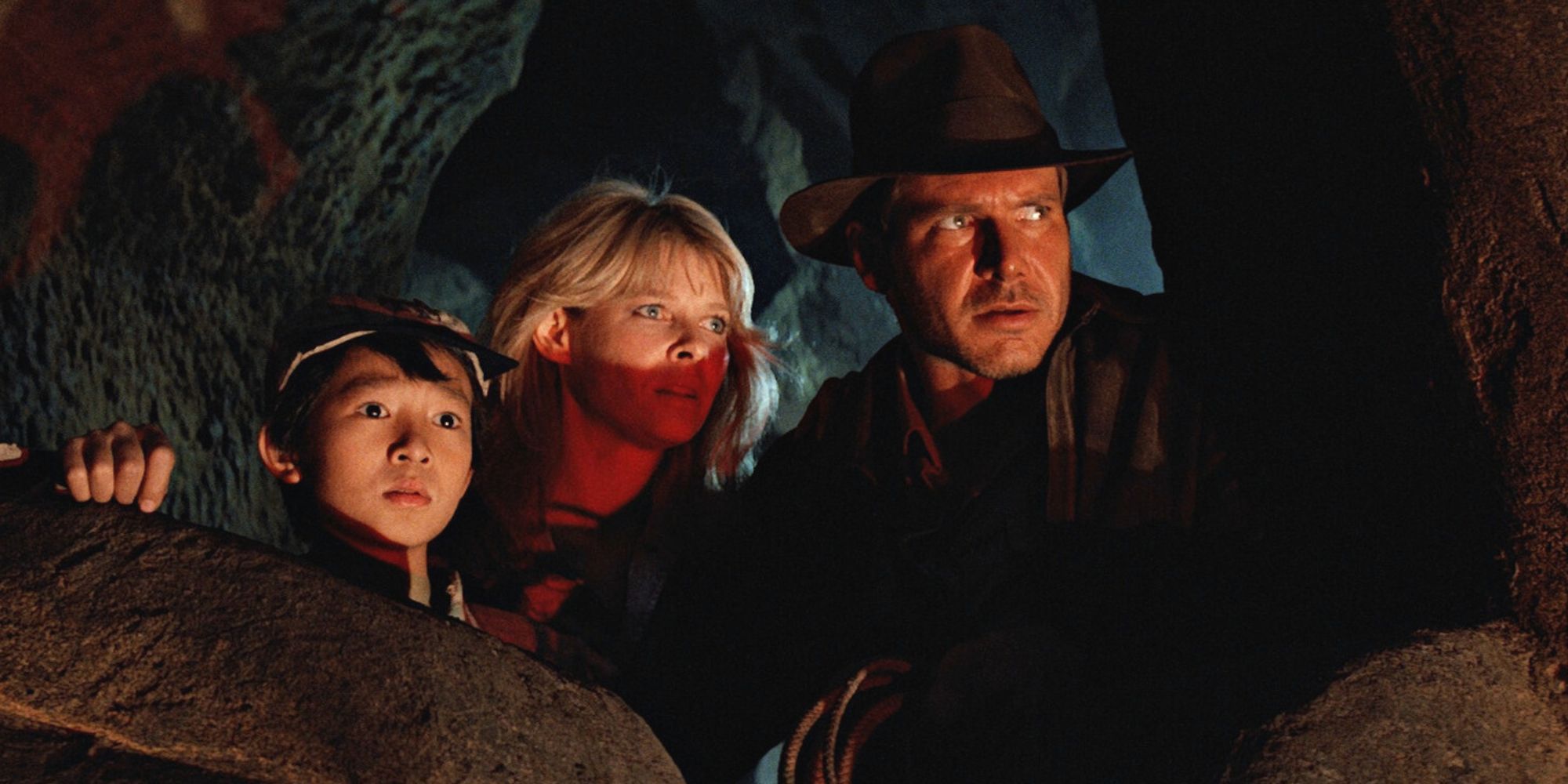 Ke Huy Quan, Kate Capshaw et Harrison Ford accroupis derrière des rochers dans une grotte regardant légèrement vers le haut dans Indiana Jones et le Temple maudit