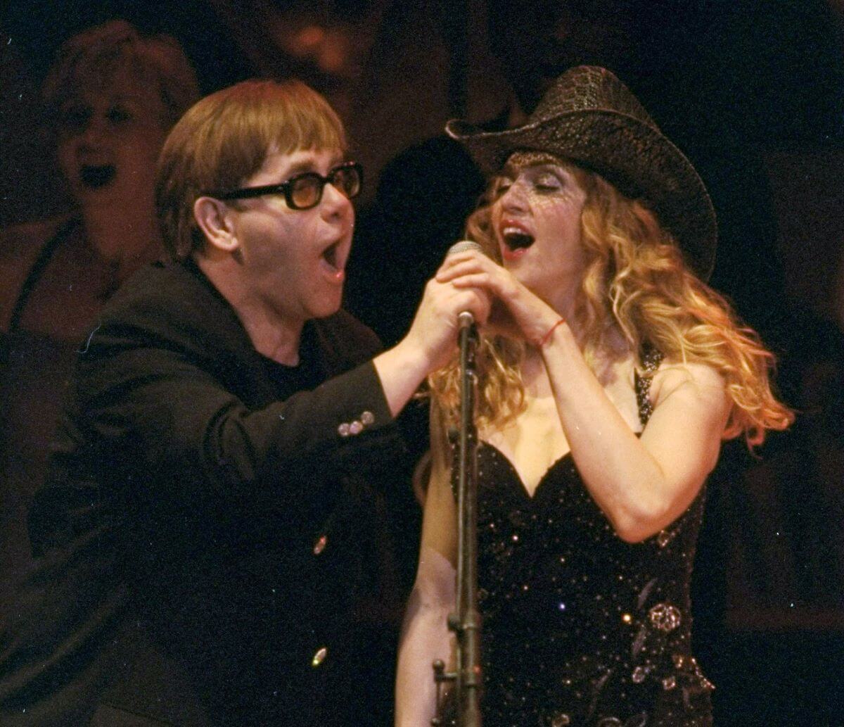 Elton John porte des lunettes de soleil et chante dans un micro avec Madonna, qui porte un chapeau de cowboy noir. Ils tiennent tous les deux une main sur le microphone.