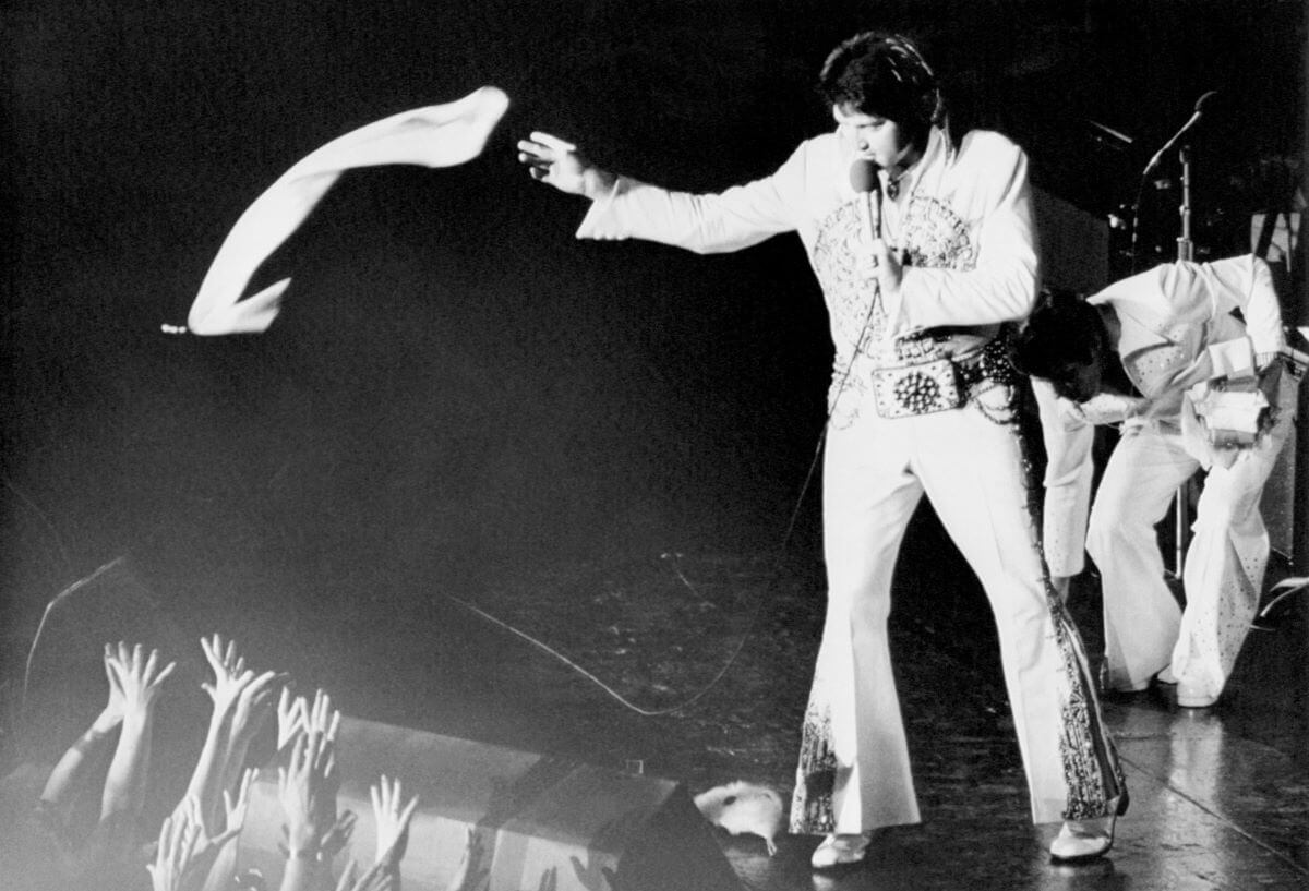 Une photo en noir et blanc d'Elvis Presley lançant un foulard à son public lors d'un concert.  Il tient un micro.