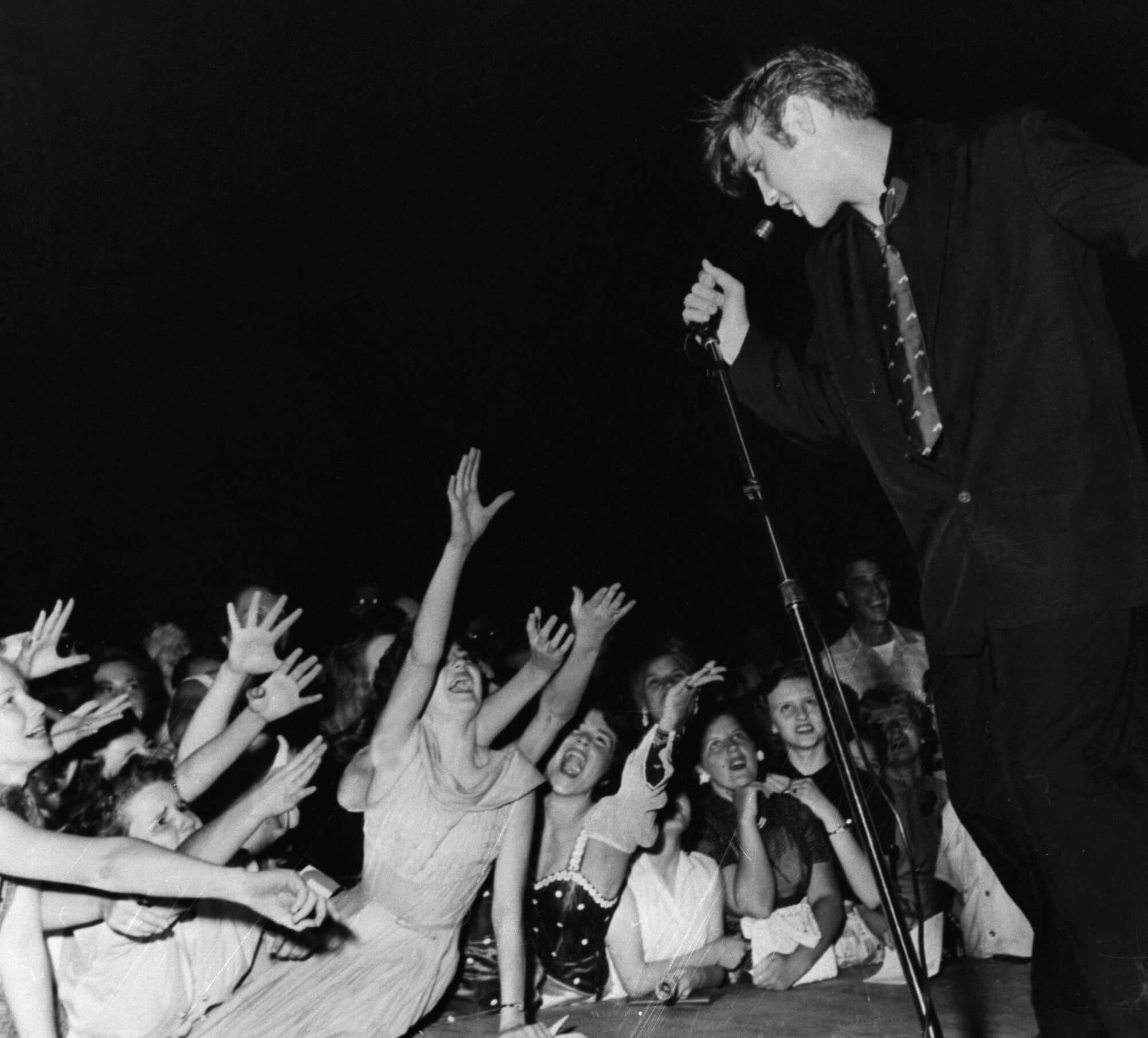 Une photo en noir et blanc d'Elvis debout sur scène et chantant devant les fans qui se tendent vers lui.