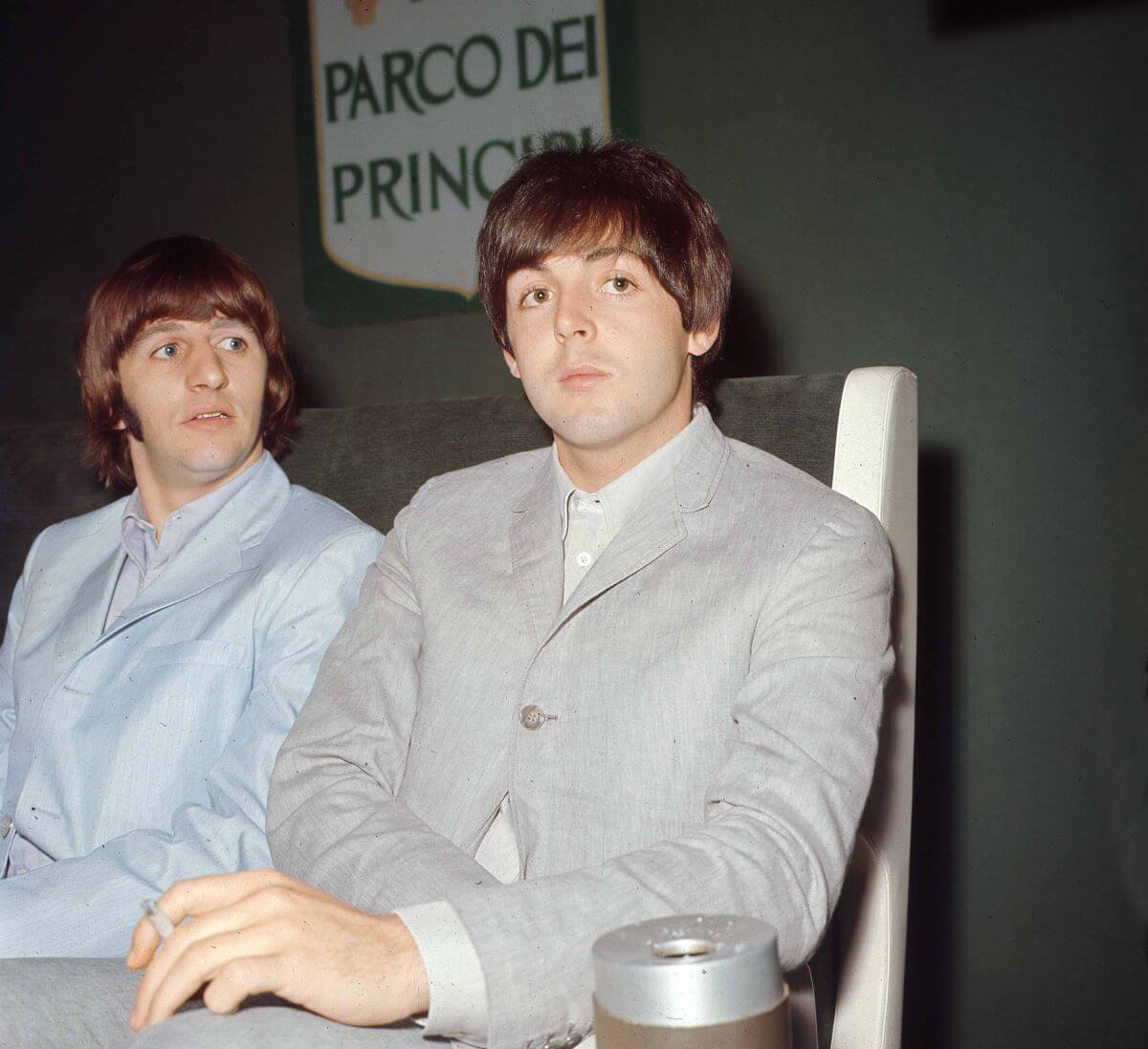 Ringo Starr et Paul McCartney sont assis ensemble du même côté d'une table. Ils portent des costumes gris.