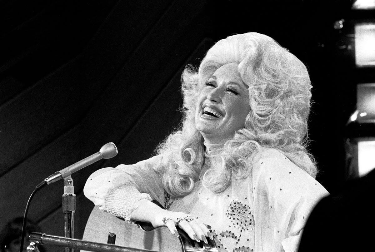 Une photo en noir et blanc de Dolly Parton posant son bras sur sa guitare et assise devant un microphone.
