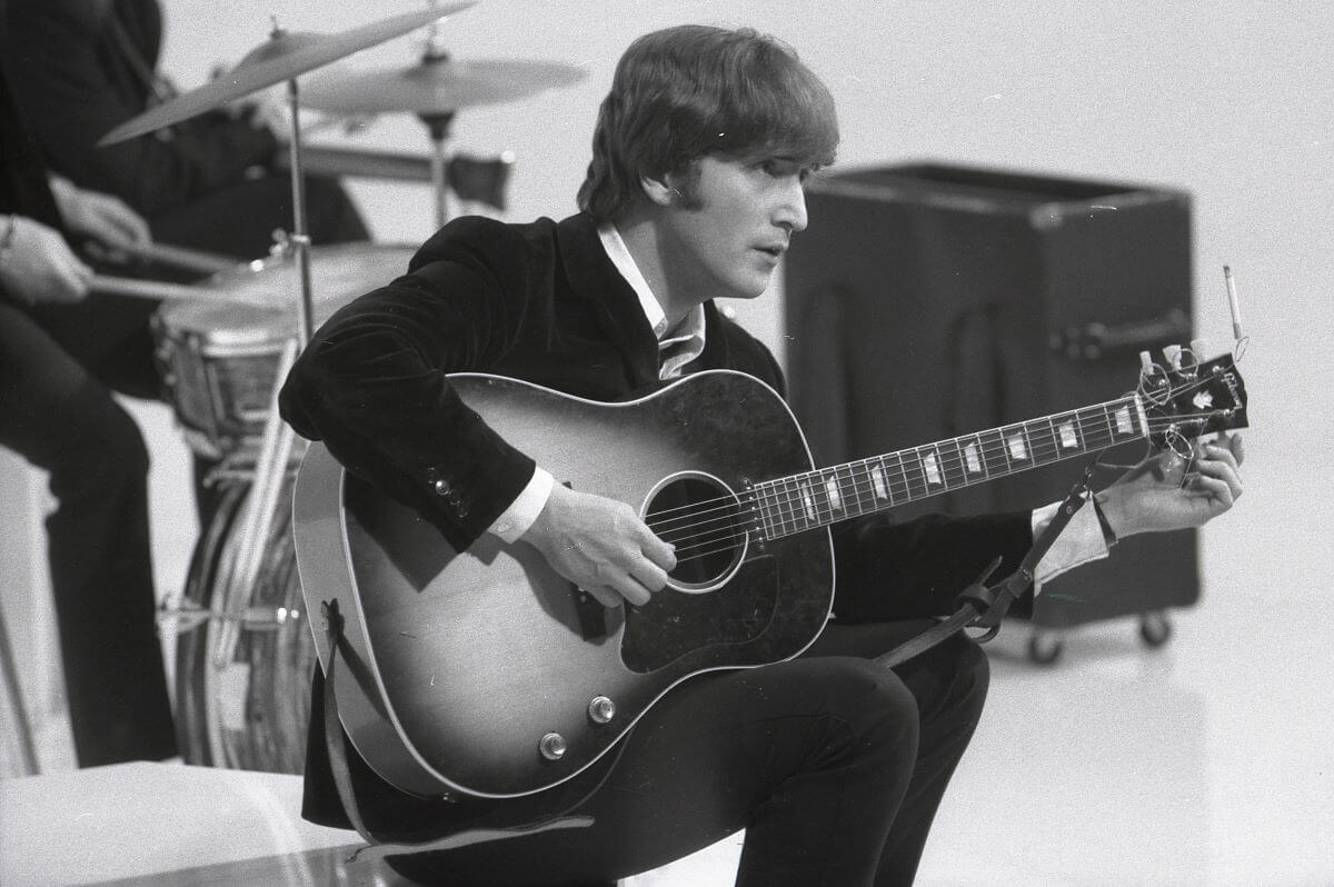 Une photo en noir et blanc de John Lennon assis et grattant une guitare acoustique.