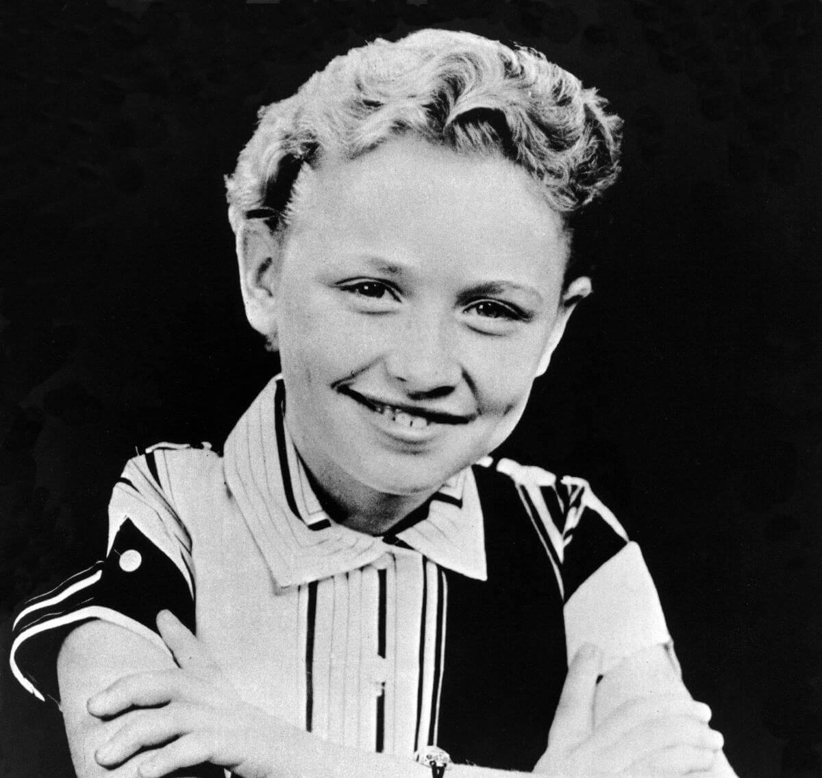 Une photo en noir et blanc de Dolly Parton enfant.  Elle porte une chemise à col et est assise les bras croisés.