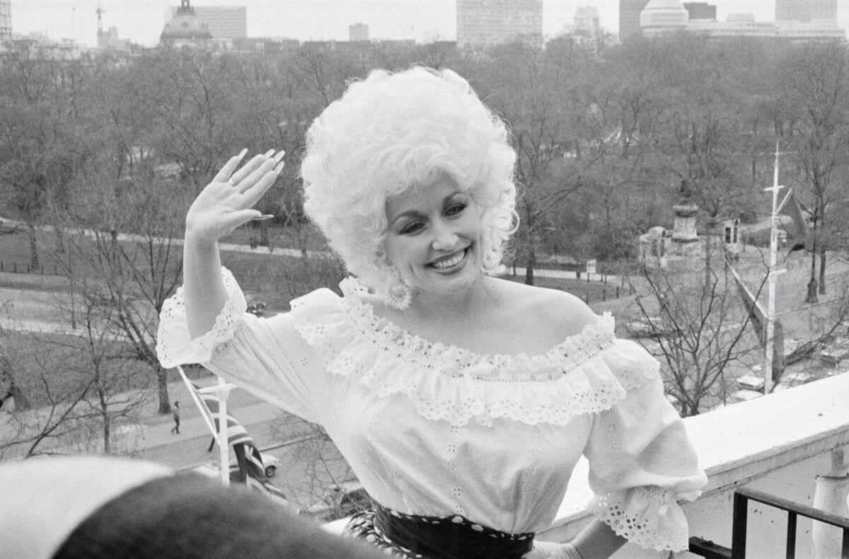 Une photo en noir et blanc de Dolly Parton debout sur un balcon et saluant.