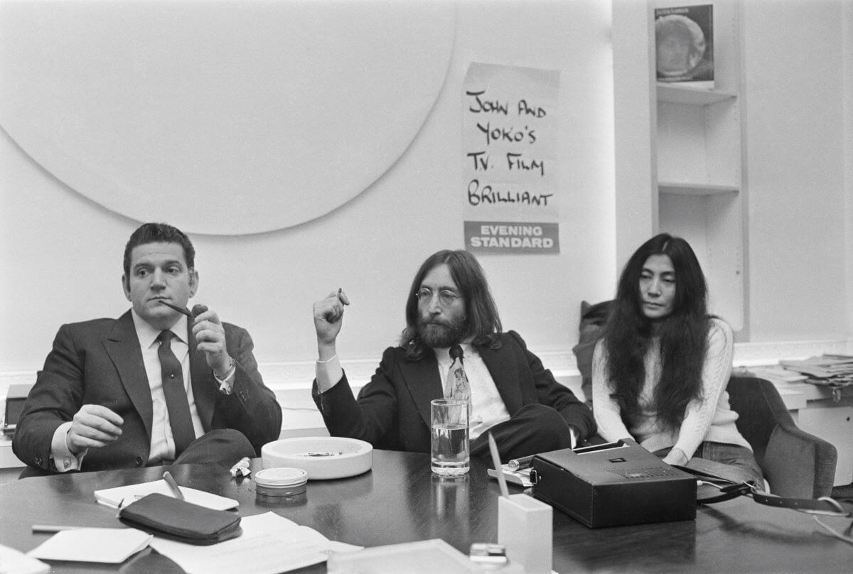 Une photo en noir et blanc d'Allen Klein, John Lennon et Yoko Ono assis d'un côté d'une table.
