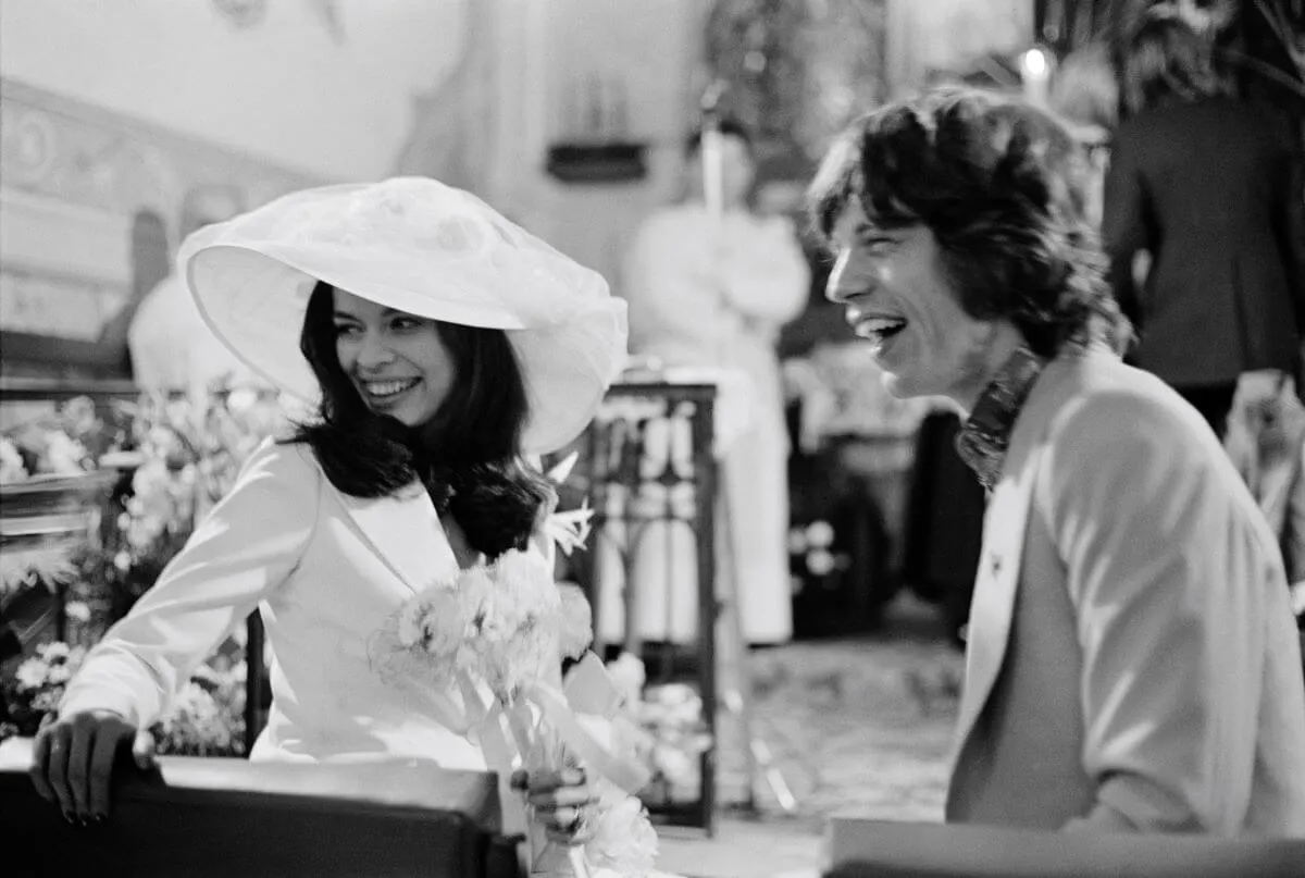 Une photo en noir et blanc de Bianca et Mick Jagger assis dans une église le jour de leur mariage.  Ils rient tous les deux.  Elle porte un chapeau et tient des fleurs.