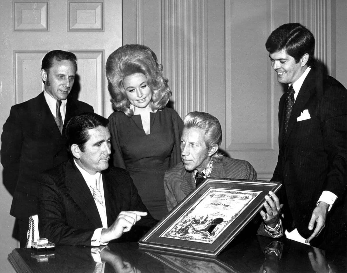 Une photo en noir et blanc de Dolly Parton et Porter Wagoner debout entre trois hommes.  Waggoner est assis et tient une plaque.