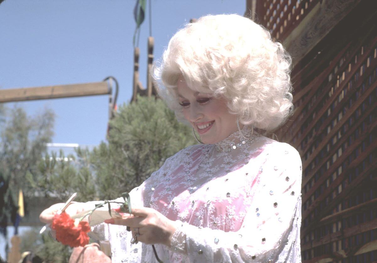 Dolly Parton porte un haut en dentelle blanche et se tient dehors.  Elle tient une fleur rouge dans une main.