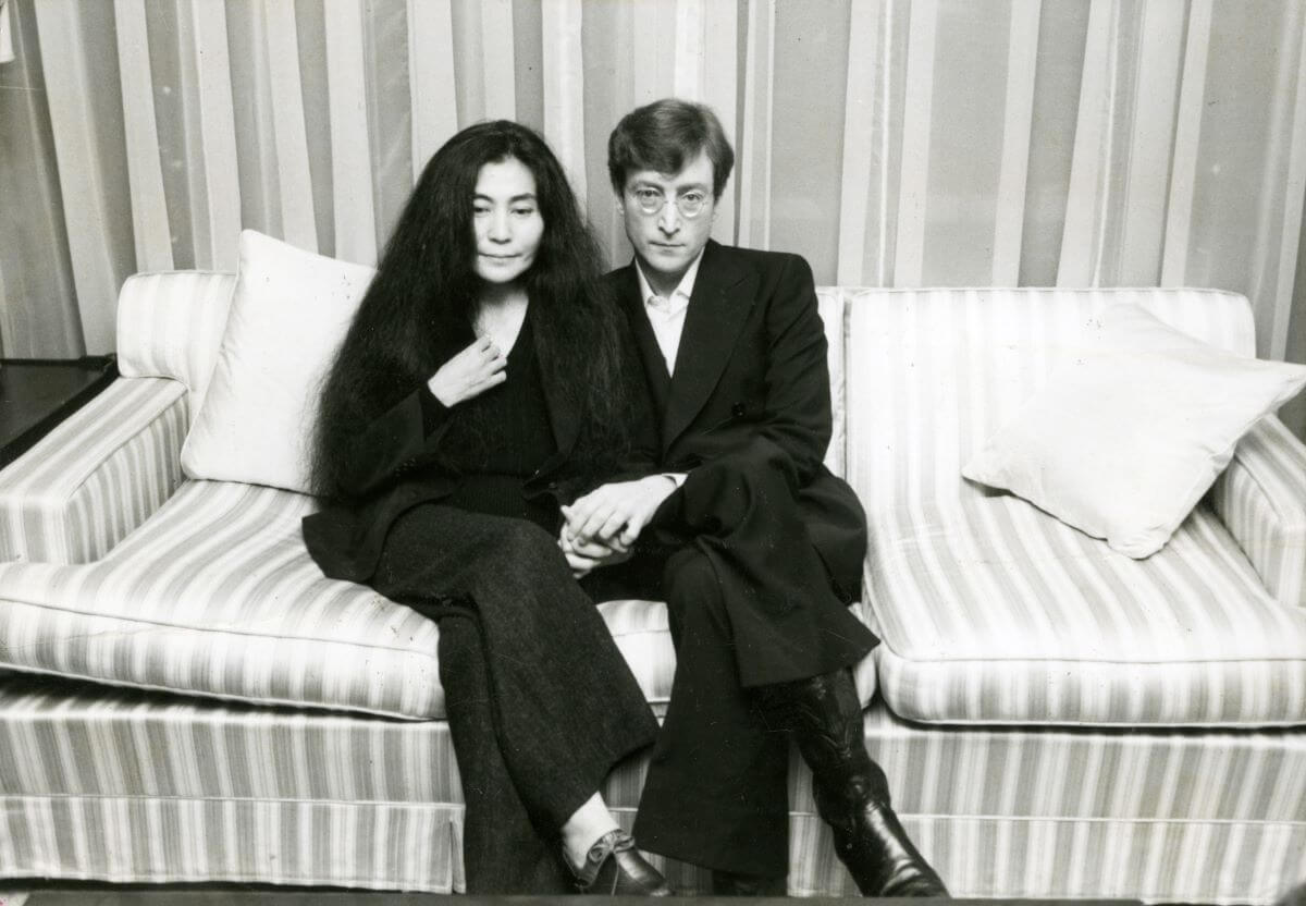 Une photo en noir et blanc de Yoko Ono et John Lennon se tenant la main et assis ensemble sur un canapé.