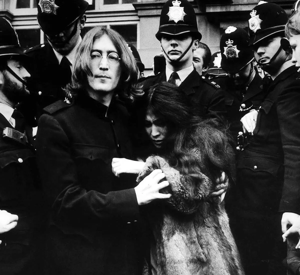Une photo en noir et blanc de John Lennon tenant Yoko Ono.  Ils sont encerclés par des policiers.