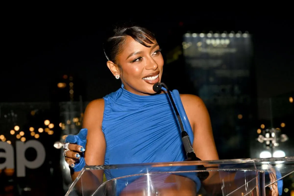 La chanteuse R&B Victoria Monet se tient sur un podium tout en portant une robe bleue.  Elle parle dans le micro.