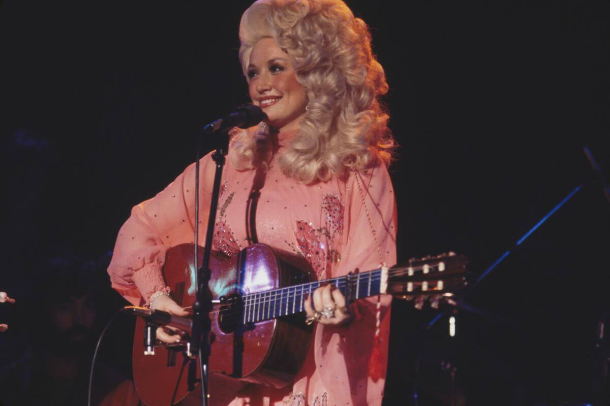 Dolly Parton porte du rose et gratte une guitare acoustique. Elle se tient devant un micro.