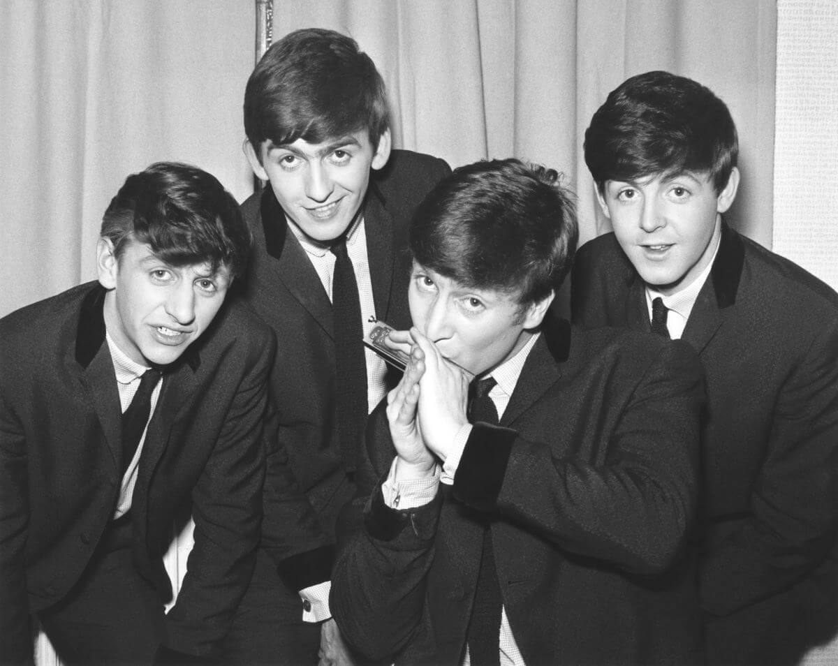 Une photo en noir et blanc des Beatles portant des costumes et se penchant les uns contre les autres.  John Lennon joue de l'harmonica.