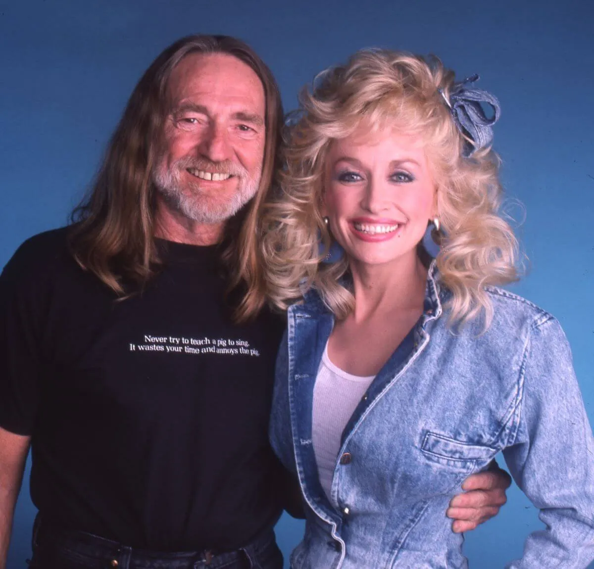 Willie Nelson porte une chemise noire et tient son bras autour de la taille de Dolly Parton.  Elle porte une veste en jean et un nœud pour cheveux en jean.
