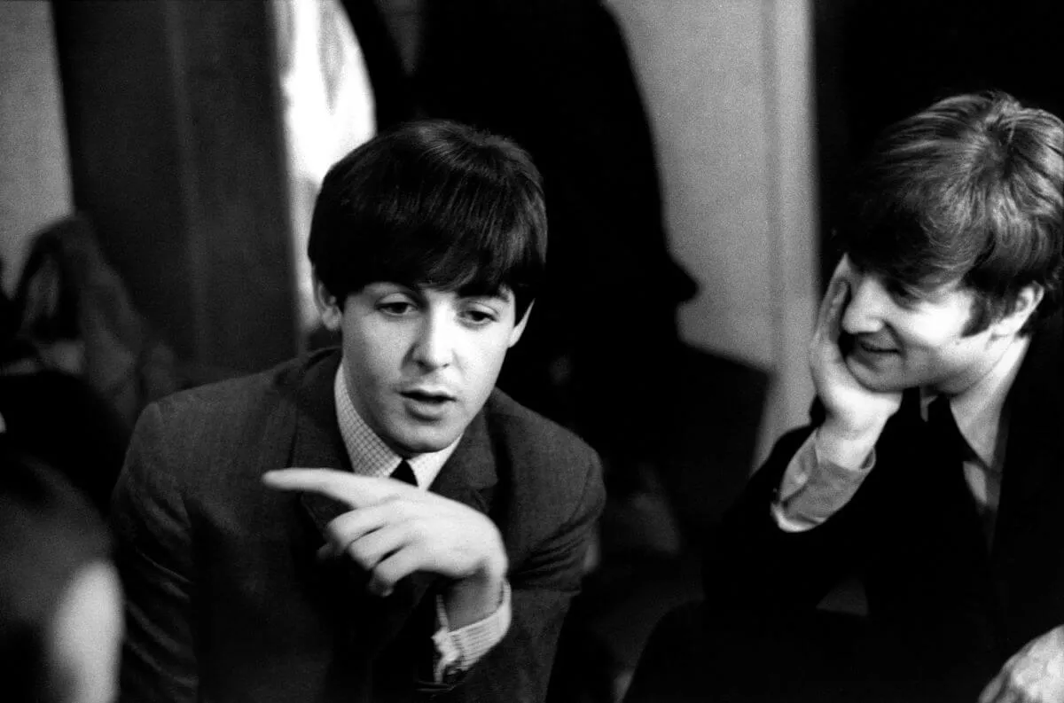 Une photo en noir et blanc de Paul McCartney et John Lennon portant des costumes.  Lennon regarde McCartney pendant qu'il parle.