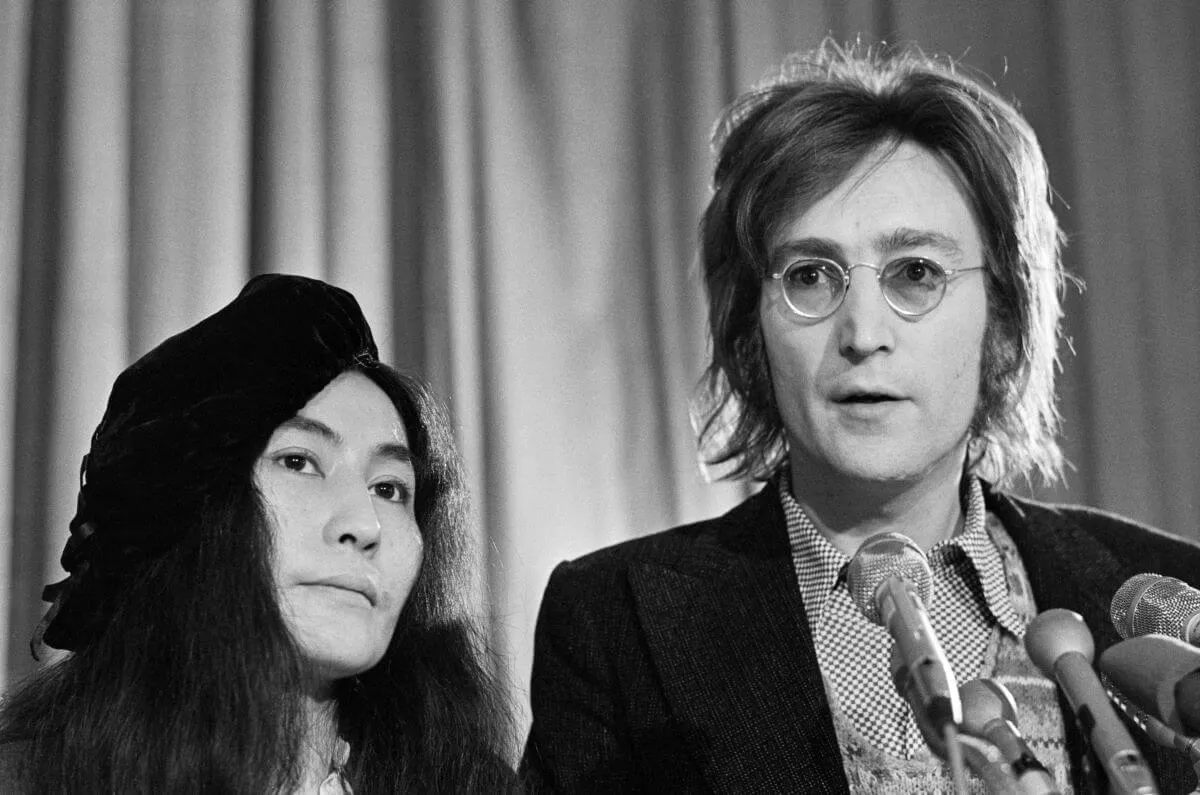 Une photo en noir et blanc de Yoko Ono debout à côté de John Lennon.  Il se tient devant plusieurs micros.