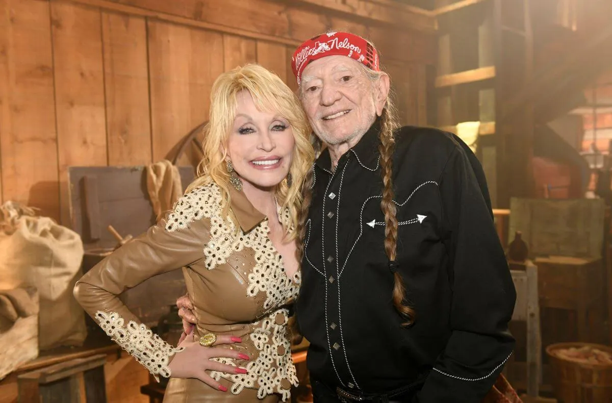 Dolly Parton et Willie Nelson se tiennent dans les bras.  Elle porte une chemise marron et blanche et il porte une chemise noire et un bandana rouge autour de la tête.