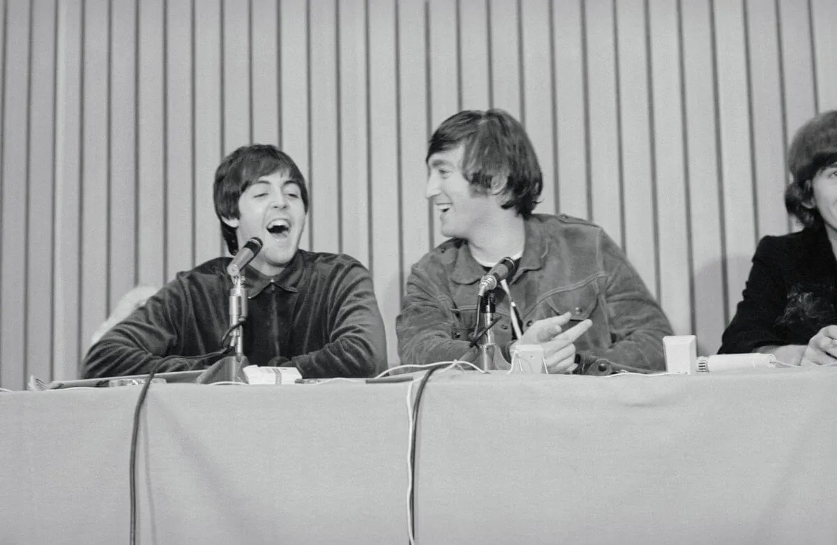 Une photo en noir et blanc de Paul McCartney et John Lennon assis derrière une table et riant.  Ils ont des micros devant eux.