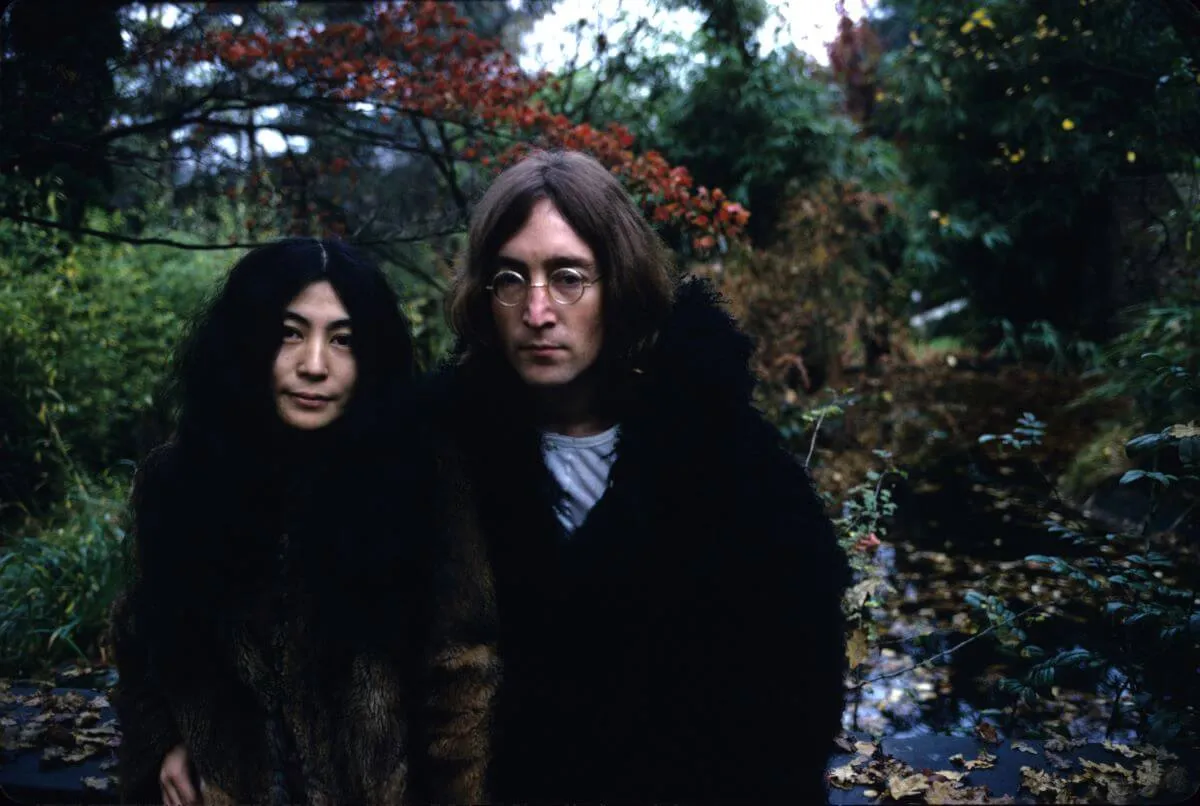 Yoko Ono et John Lennon se tiennent devant une zone boisée.  Ils portent tous les deux des manteaux.