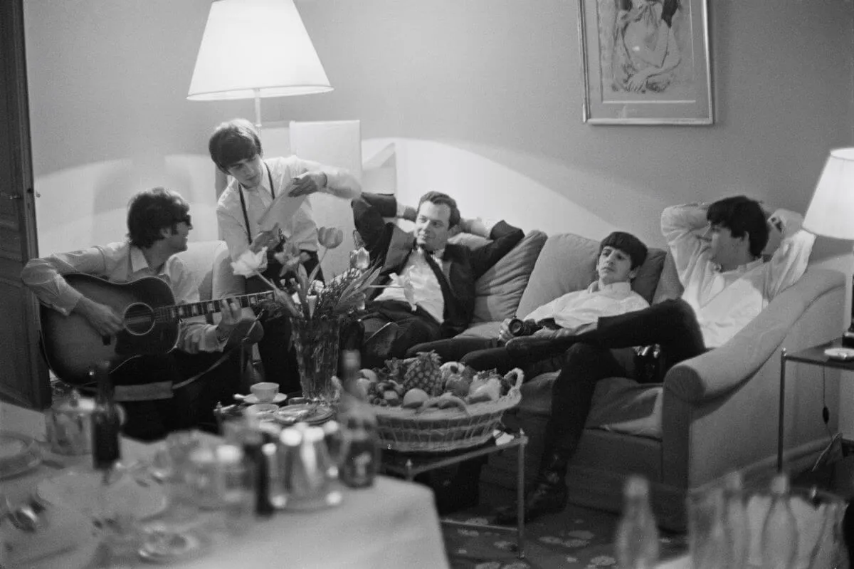 Une photo en noir et blanc de Brian Epstein assis entre les Beatles sur un canapé. Ils se détendent tous sauf John Lennon, qui joue de la guitare.