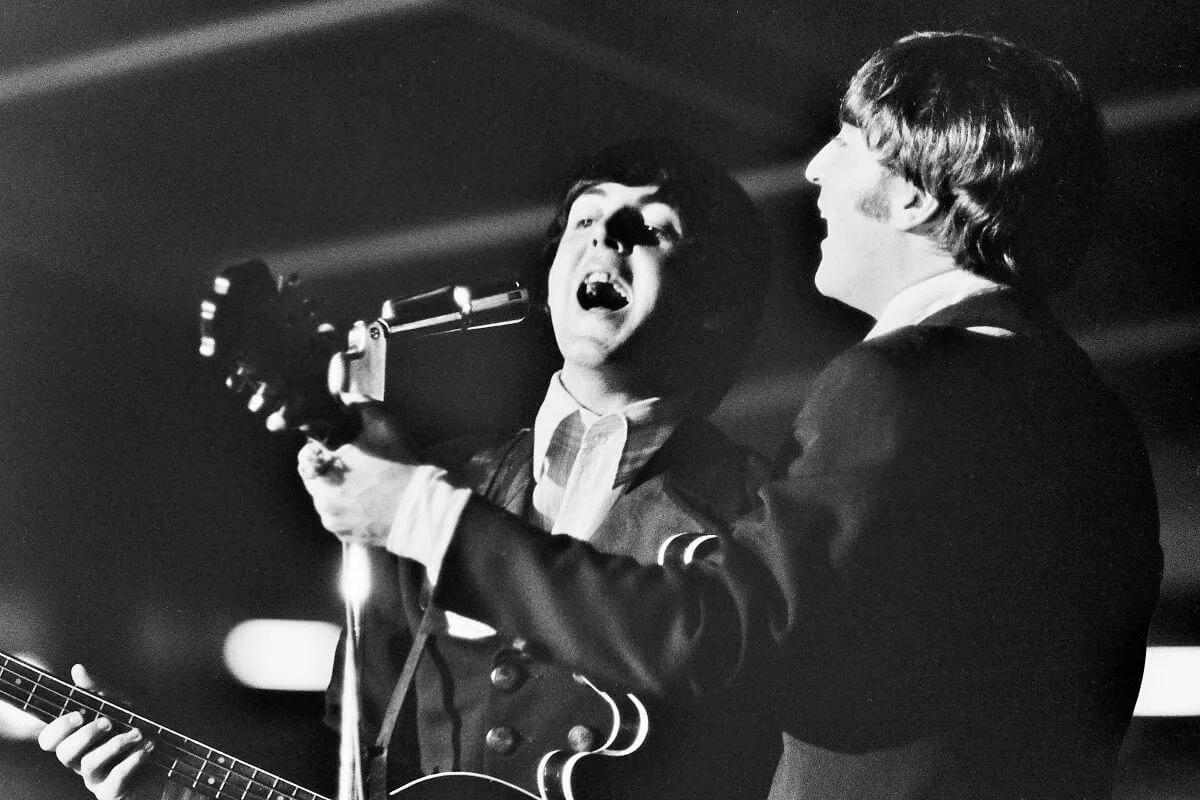Une photo en noir et blanc de Paul McCartney et John Lennon chantant dans le même micro. Lennon tourne le dos à la caméra et McCartney est orienté vers elle.