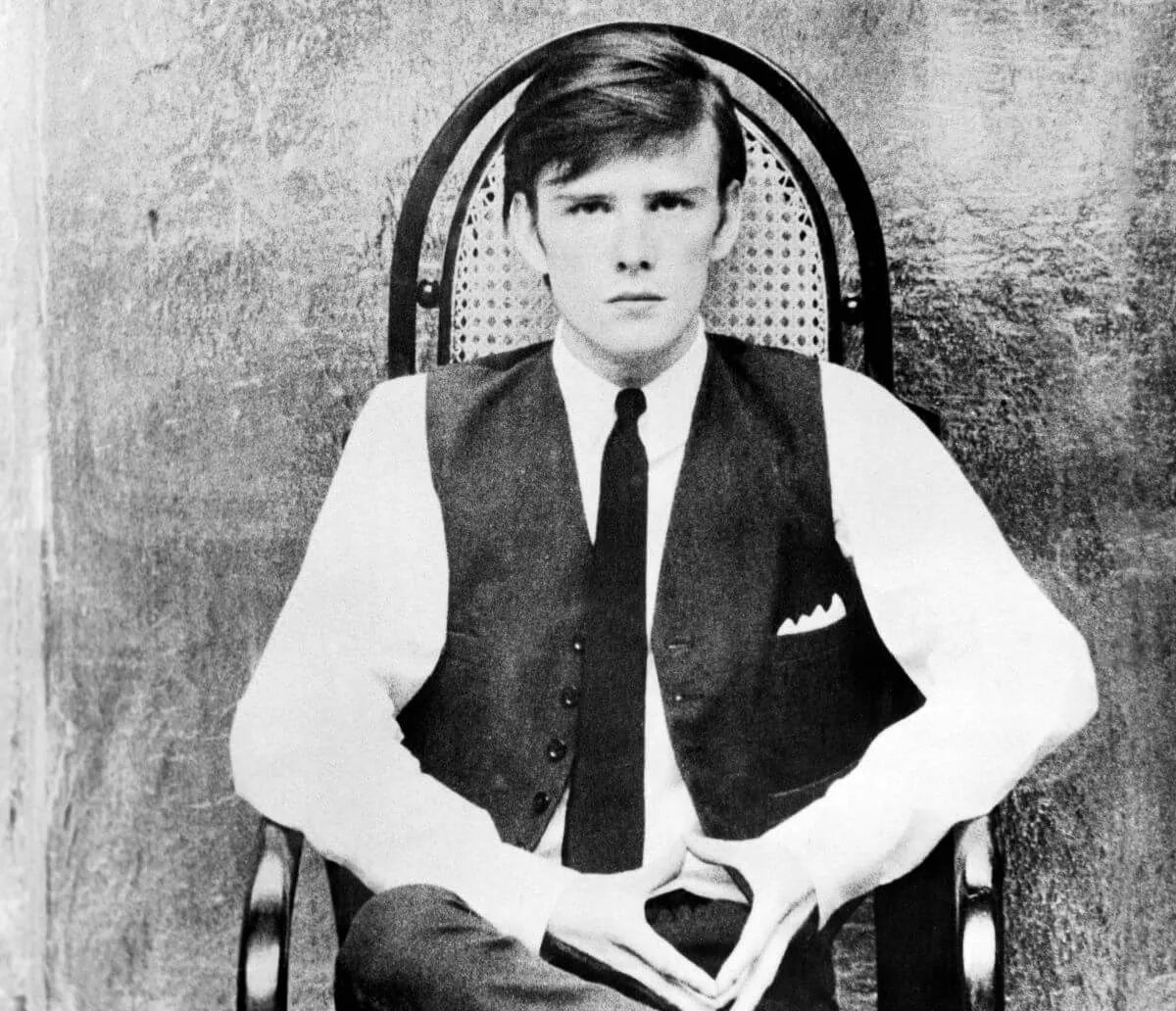 Une photo en noir et blanc de Stuart Sutcliffe portant une cravate et assis sur une chaise.