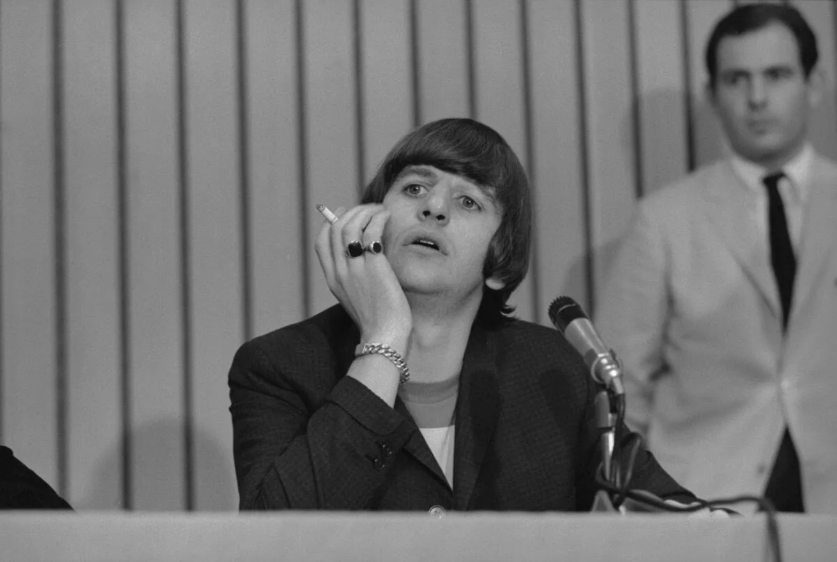 Une photo en noir et blanc de Ringo Starr assis devant un micro, le menton posé sur sa main.  Il tient une cigarette entre ses doigts.
