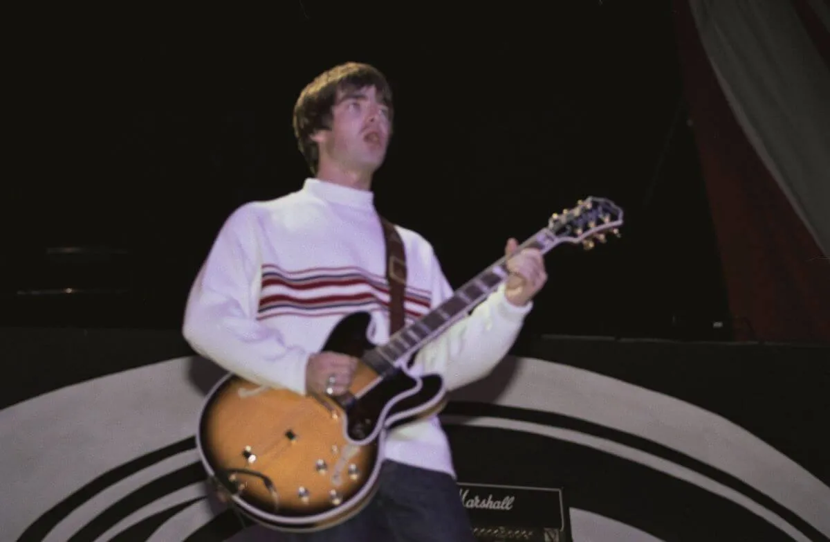 Noel Gallagher porte une chemise blanche et joue de la guitare.