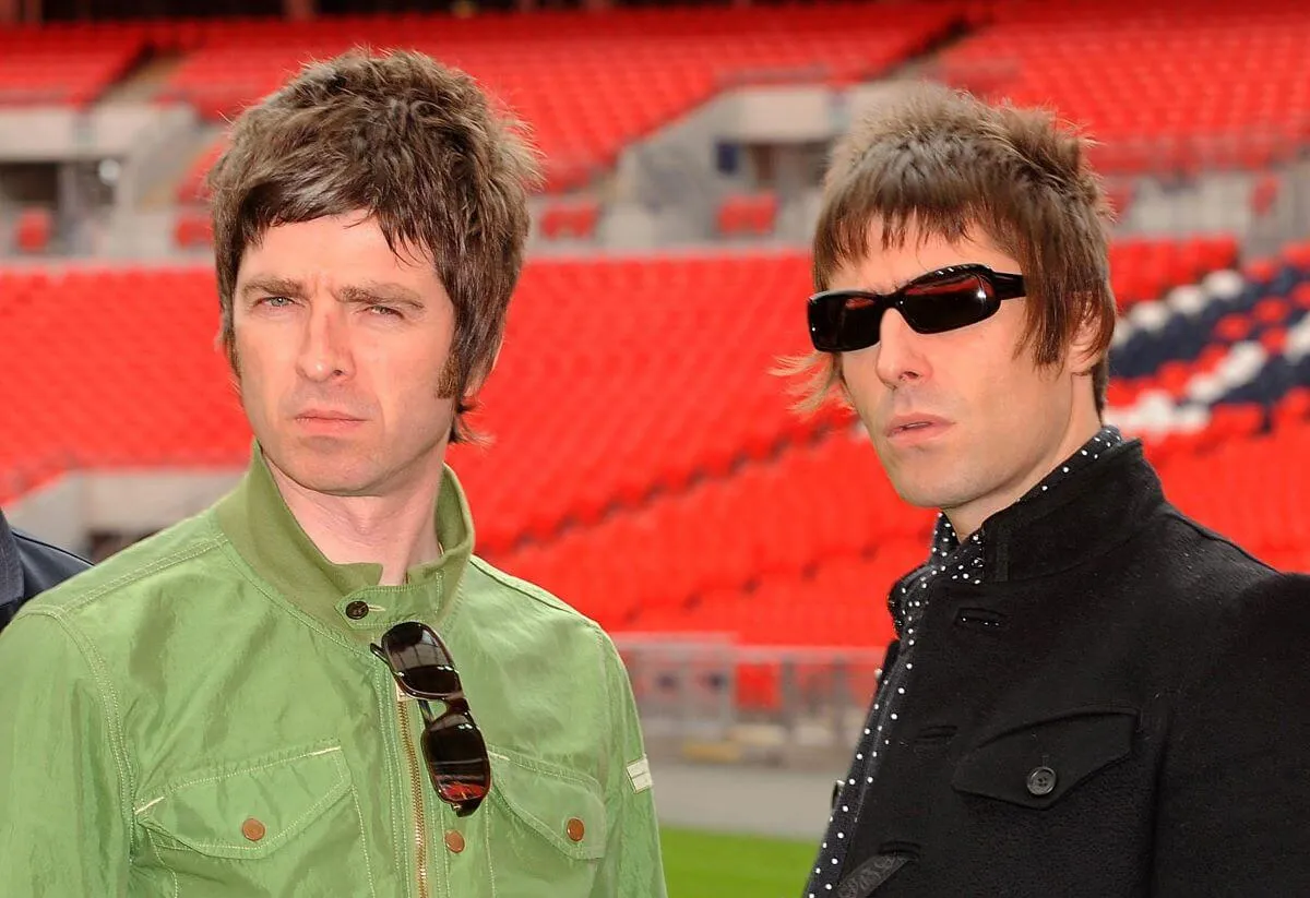 Noel Gallagher porte un manteau vert et se tient à côté de Liam Gallagher, qui porte un manteau noir. Ils se tiennent dans un stade de football.
