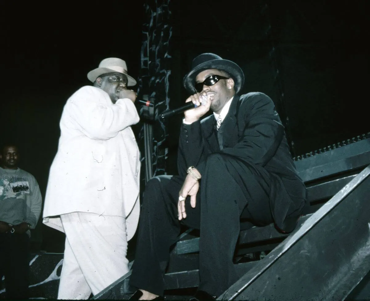Le Notorious BIG porte un costume et un chapeau blancs et tient un microphone. Sean « Diddy » Combs porte un costume et un chapeau noirs et est assis tout en tenant un microphone.