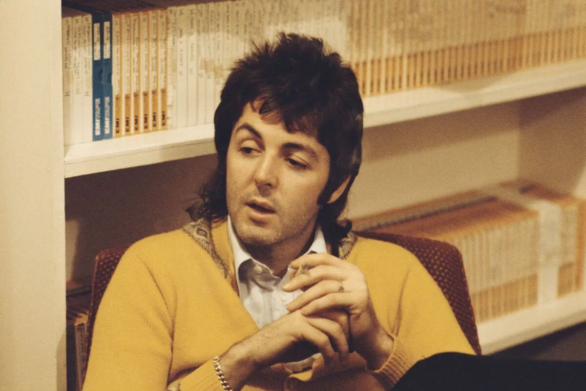 Le musicien britannique Paul McCartney porte un pull jaune et est assis devant des étagères.