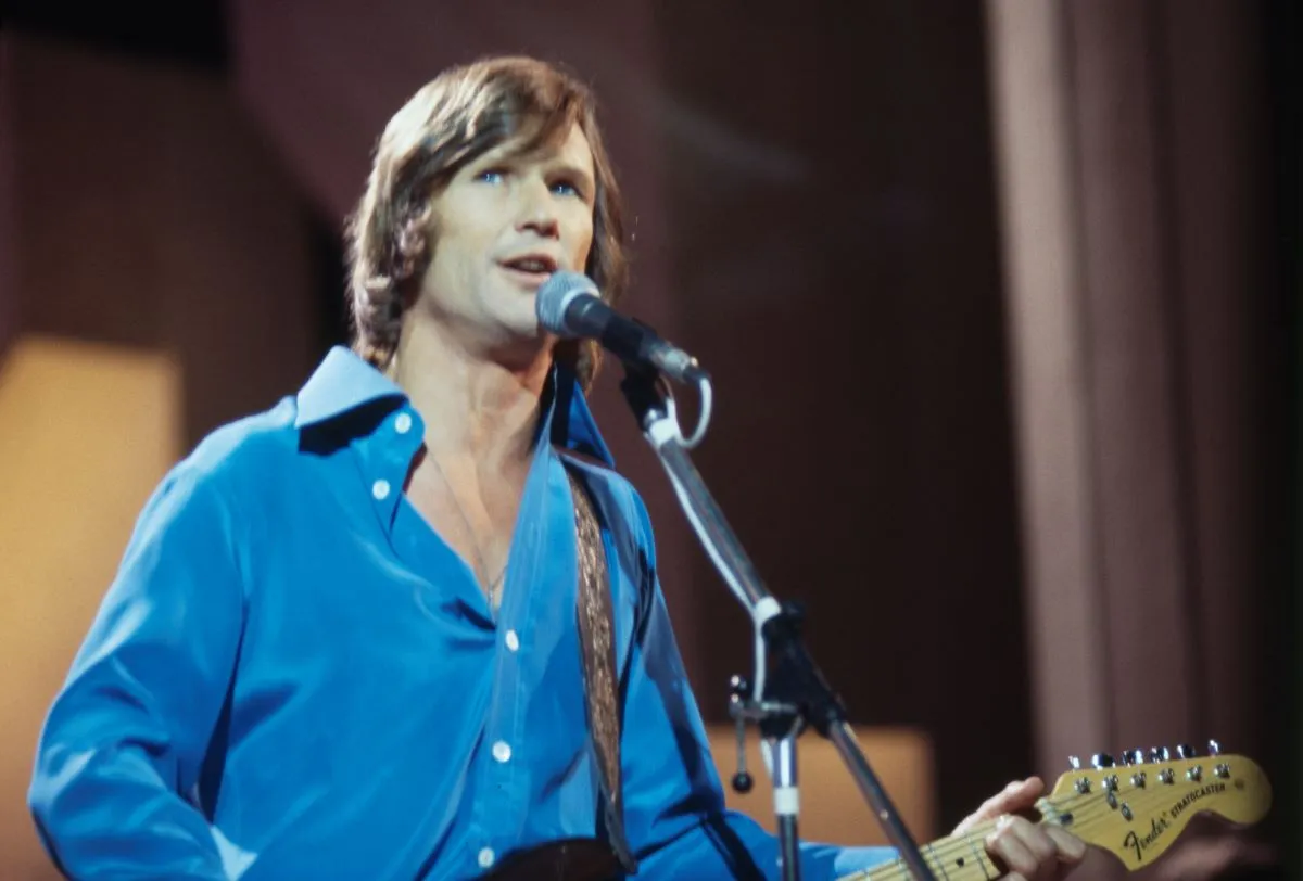 Kris Kristofferson porte une chemise bleue et chante dans un micro.