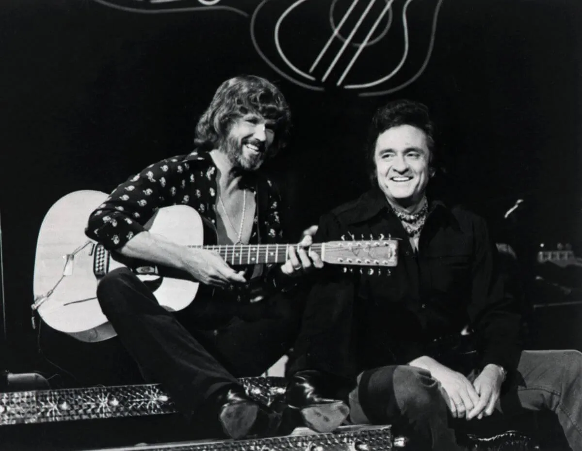 Une photo en noir et blanc de Kris Kristofferson et Johnny Cash assis ensemble. Kristofferson tient une guitare.