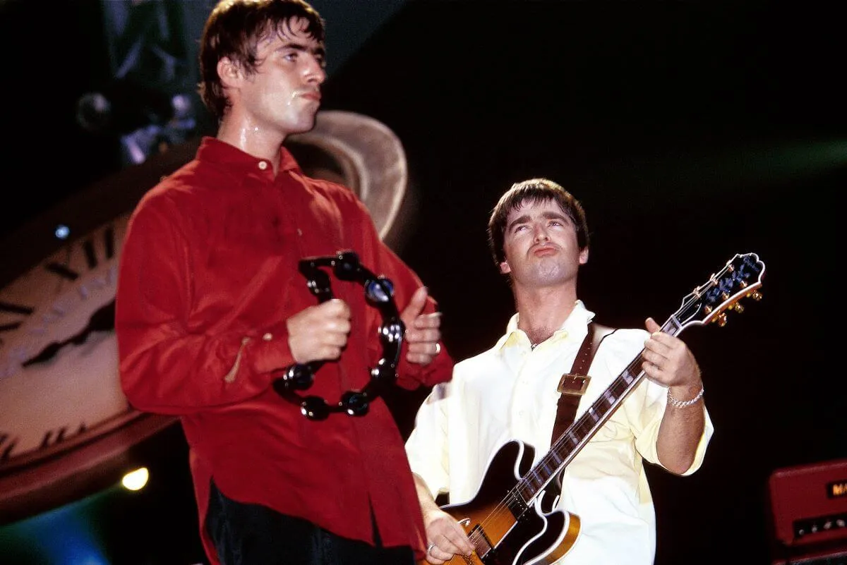 Liam et Noel Gallagher d'Oasis sont sur scène. Liam porte une chemise rouge et joue du tambourin. Noel porte une chemise blanche et joue de la guitare.