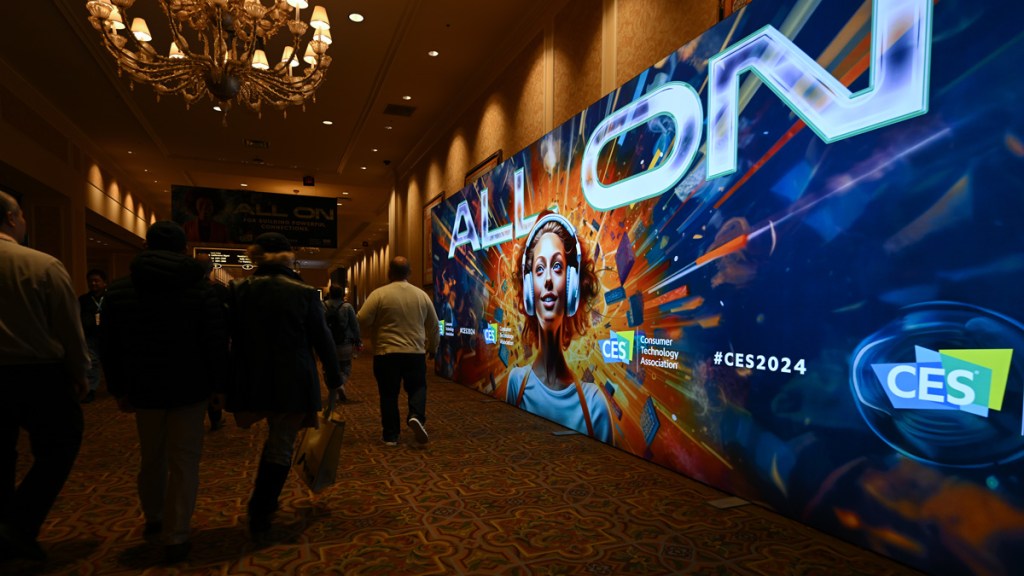 Les participants au CES 2024 passent devant un grand panneau d'affichage numérique qui indique "TOUT ON" sur une photo d’une femme avec des écouteurs. Explosion de feu derrière sa tête.