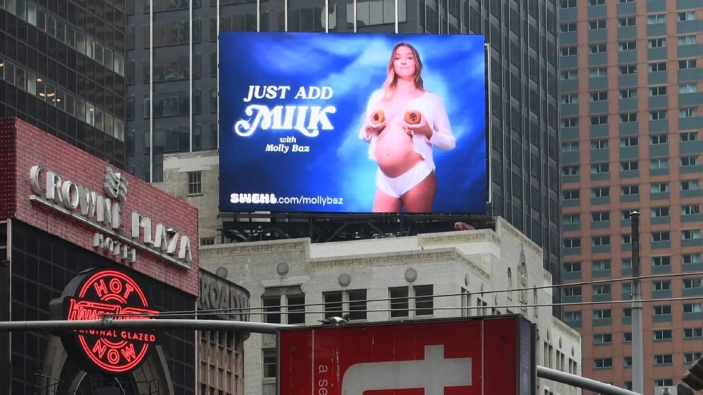 Panneau d'affichage numérique bleu vif à Times Square montrant une femme tenant des biscuits devant ses seins tout en portant une chemise blanche boutonnée et des sous-vêtements. Le texte dit